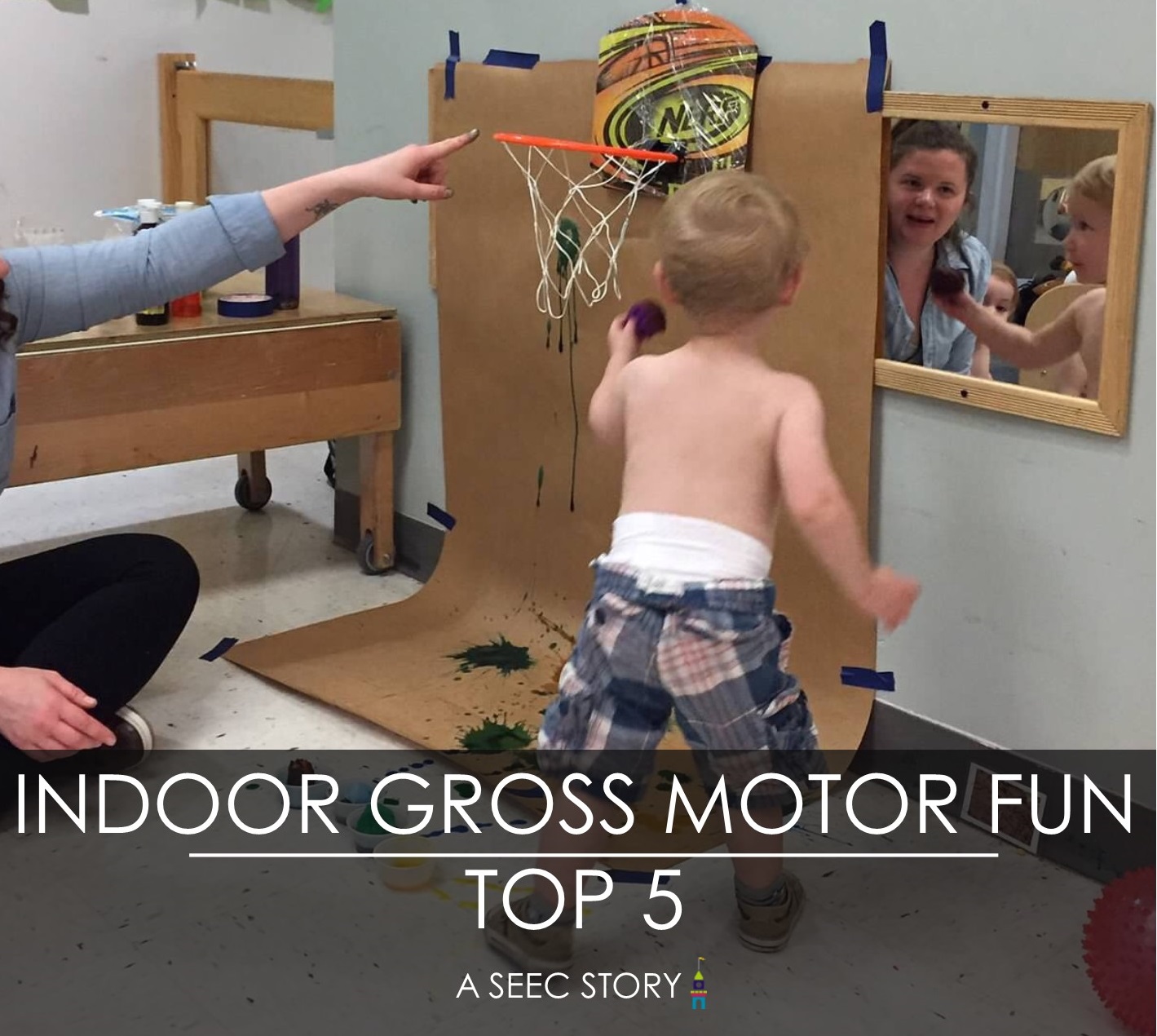 A toddler holds a pom pom with paint on it in front of a little basketball hoop with butcher paper behind it. The butcher paper has paint on it. An adult is behind the child pointing at the basketball hoop.