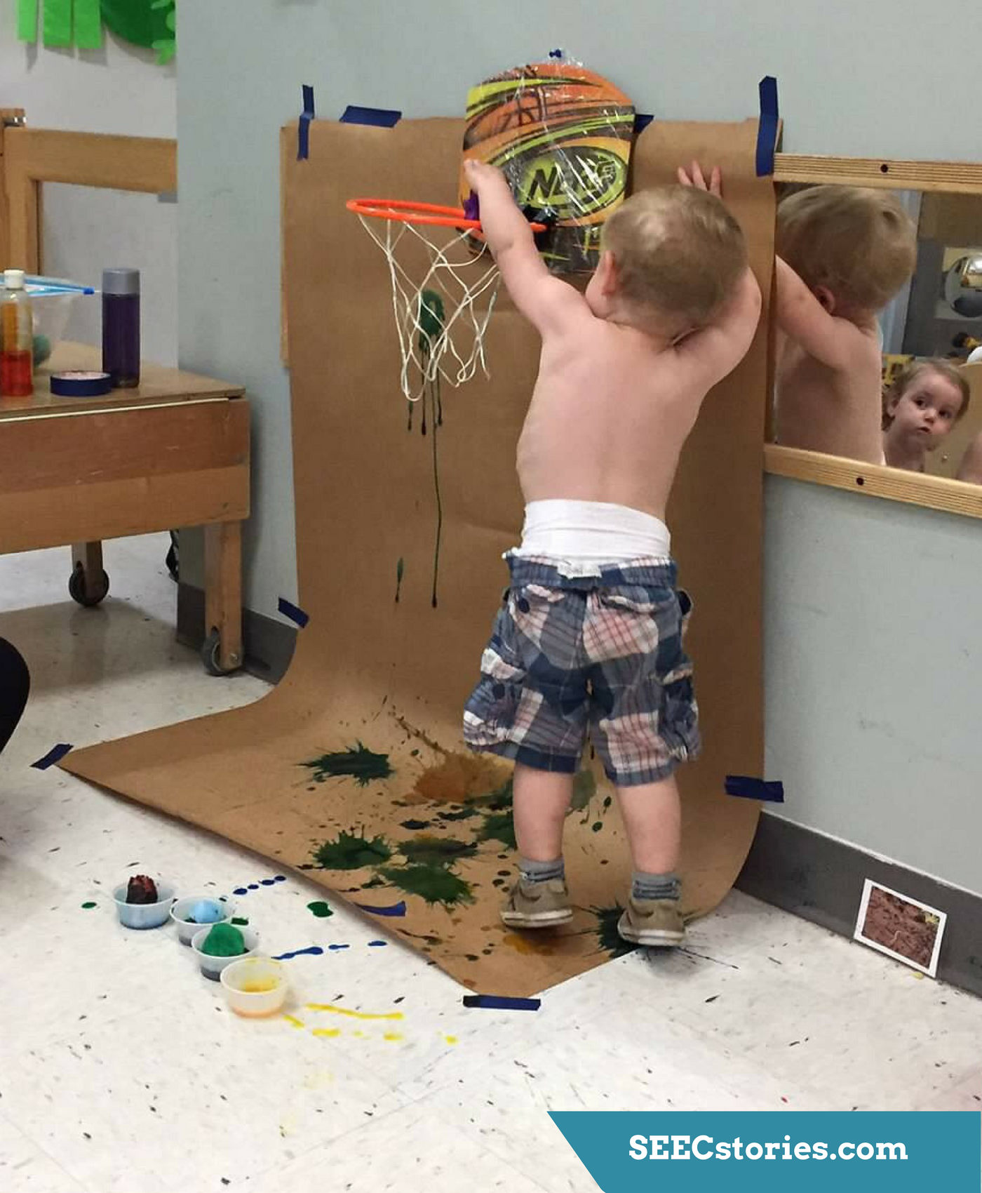 A toddler dunks a pom pom with paint on it into a little basketball hoop with butcher paper behind it. The butcher paper has paint on it.